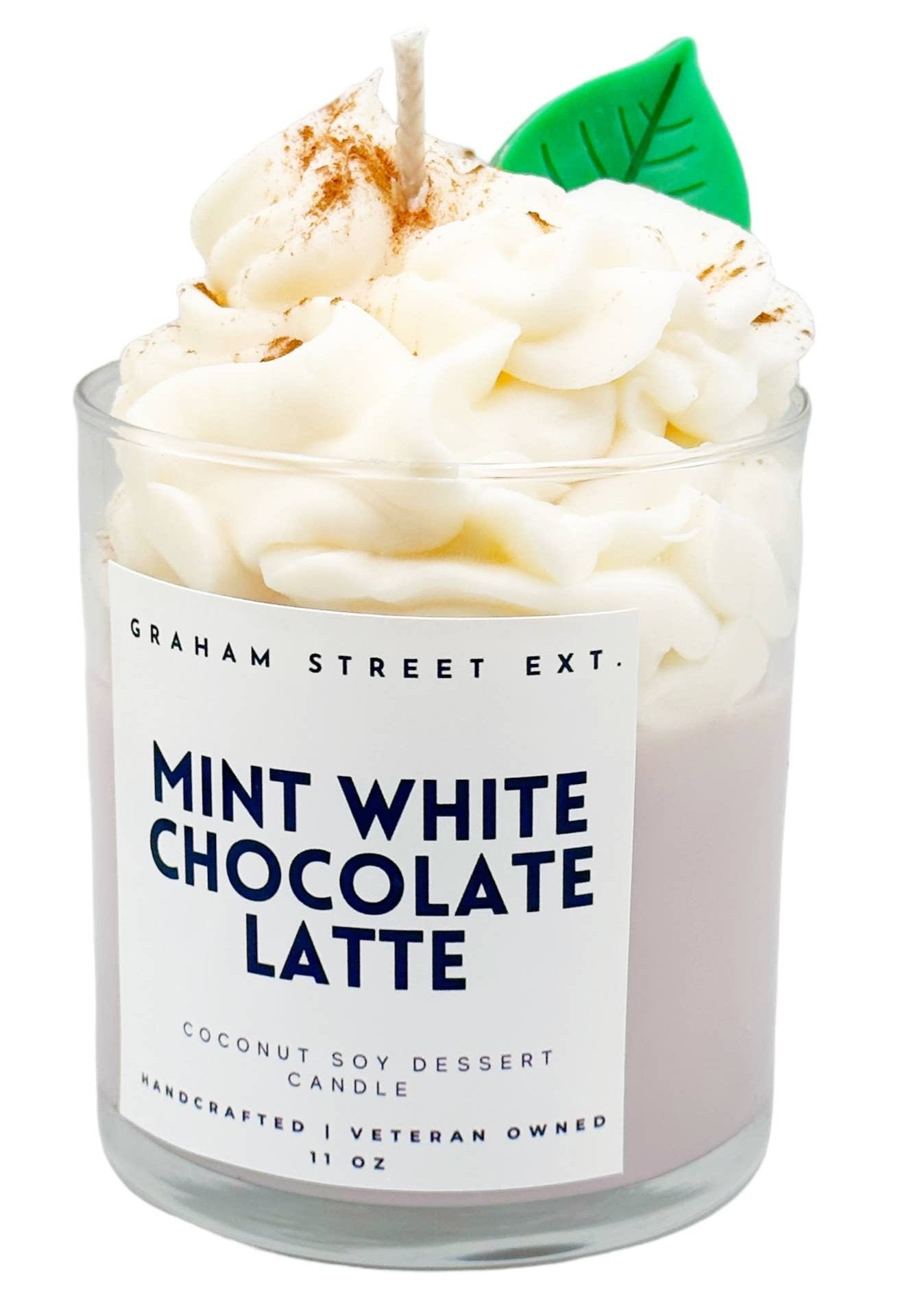 A handcrafted dessert-style candle designed to look like a mint white chocolate latte. The candle is housed in a clear glass jar labeled with "Graham Street Ext. MINT WHITE CHOCOLATE LATTE" and described as a "Coconut Soy Dessert Candle." The realistic whipped cream topping, made of wax, is sprinkled with brown accents resembling cinnamon and features a small green wax leaf for added detail. This 11 oz candle, handcrafted by a veteran-owned business, combines visual charm with a cozy aesthetic.