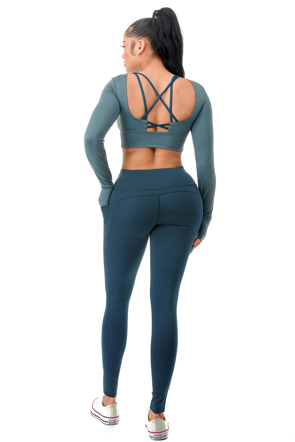 A model wearing a stylish teal two-piece activewear set consisting of a long-sleeve cropped top and high-waisted leggings. The top features a fitted design with thumbholes and a crisscross strap detail at the open back, providing both support and style. The leggings have a smooth, high-rise waistband with a seamless look, complementing the fitted silhouette. The model completes the outfit with white sneakers and a high ponytail, showcasing a sporty and chic appearance. Perfect for workouts, yoga,