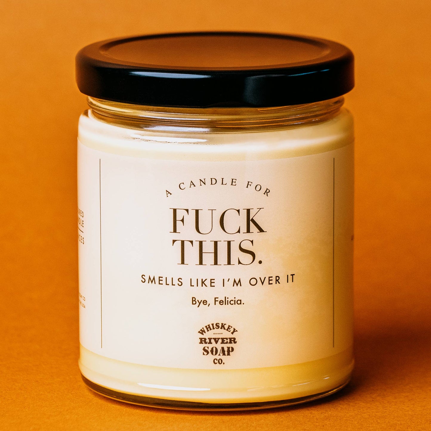 A creamy white jar candle by Whiskey River Soap Co. with bold black text reading, "A Candle for F*** This: Smells like I’m over it. Bye, Felicia." The candle features a sleek black lid and is set against a warm orange background, embodying humor and sarcasm for those who’ve had enough.