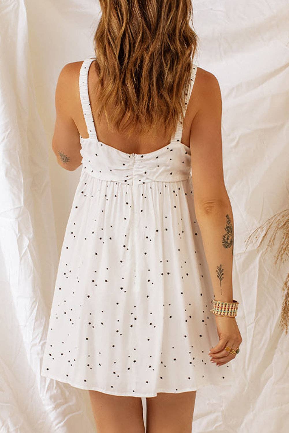 A woman wearing a white sundress with thin straps, ruched detailing at the bust, and a black polka dot pattern throughout. The dress flares out slightly, giving it a flowy look. She accessorizes with layered gold necklaces, a bracelet, and has tattoos on her arm, including a palm tree design. The background features soft white fabric and dried grasses, adding a bohemian vibe.