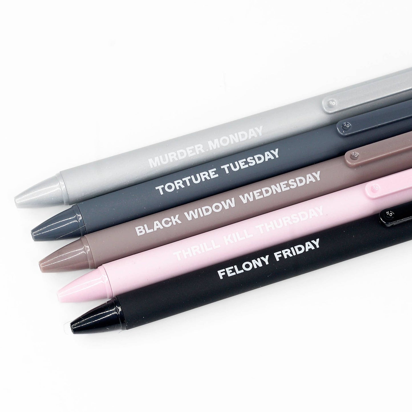 Monochrome pen set in sleek gray, black, and blush tones with phrases like 'Murder Monday' and 'Felony Friday,' packaged in a pink box. A quirky and edgy addition to any desk or writing set.