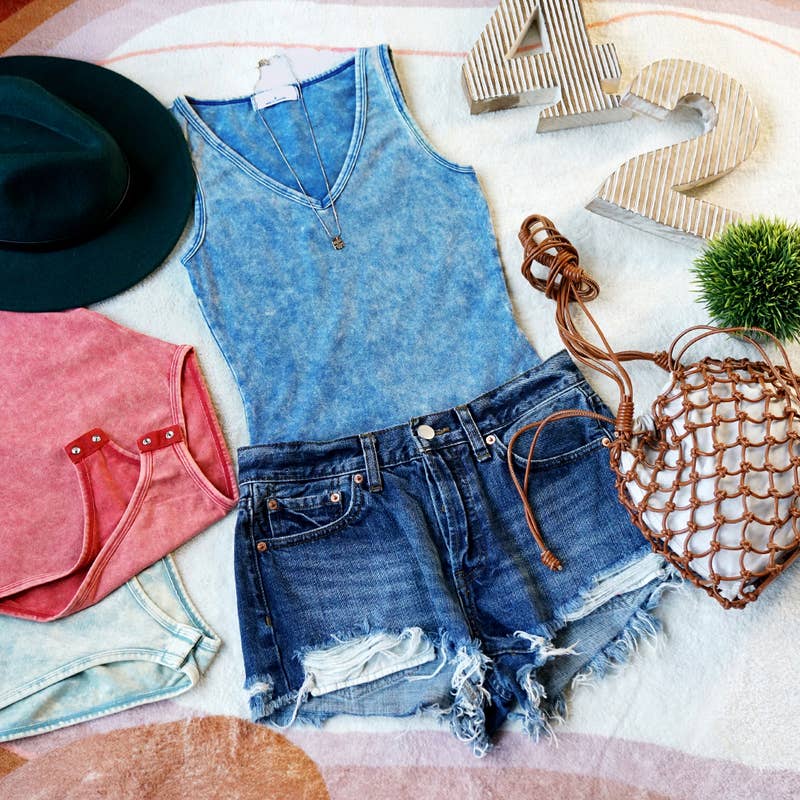Flat lay of a casual summer outfit featuring a light blue acid-wash tank top, distressed denim shorts, a green hat, a brown macrame bag, and pink and aqua tank tops in the background.