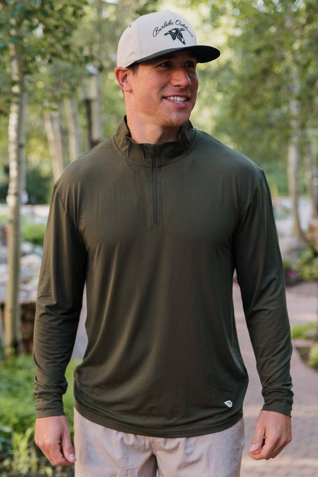 Man wearing a burlebo olive-green long-sleeve quarter-zip pullover shirt with a light gray and black logo cap, standing outdoors surrounded by trees.