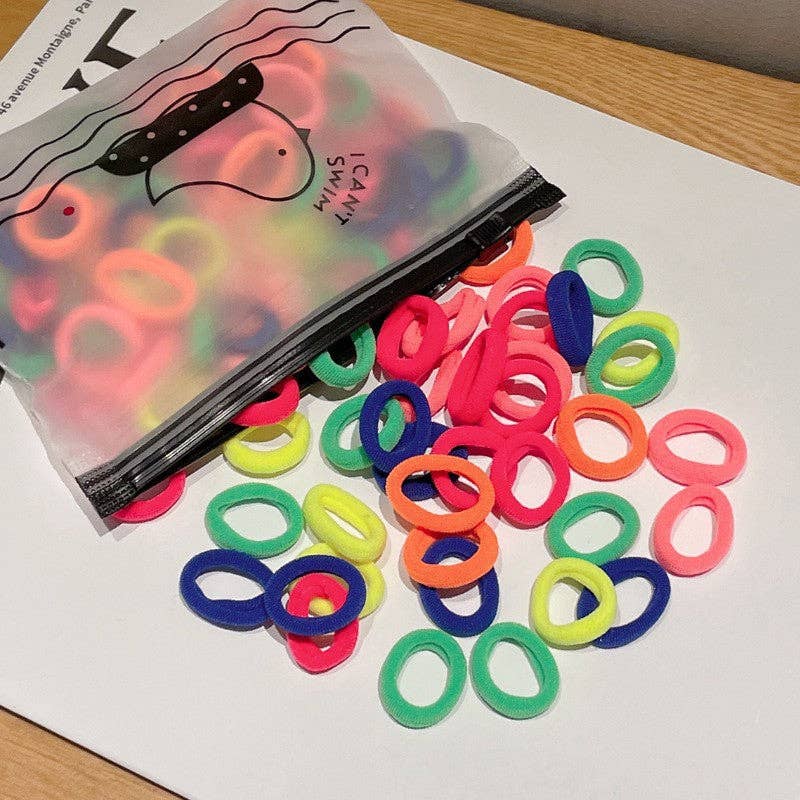 A small plastic zippered bag labeled "I Can Swim" is open, spilling out a colorful assortment of hair ties onto a white surface. The hair ties come in soft pastel shades, including pink, purple, yellow, green, and blue, creating a vibrant and playful display.