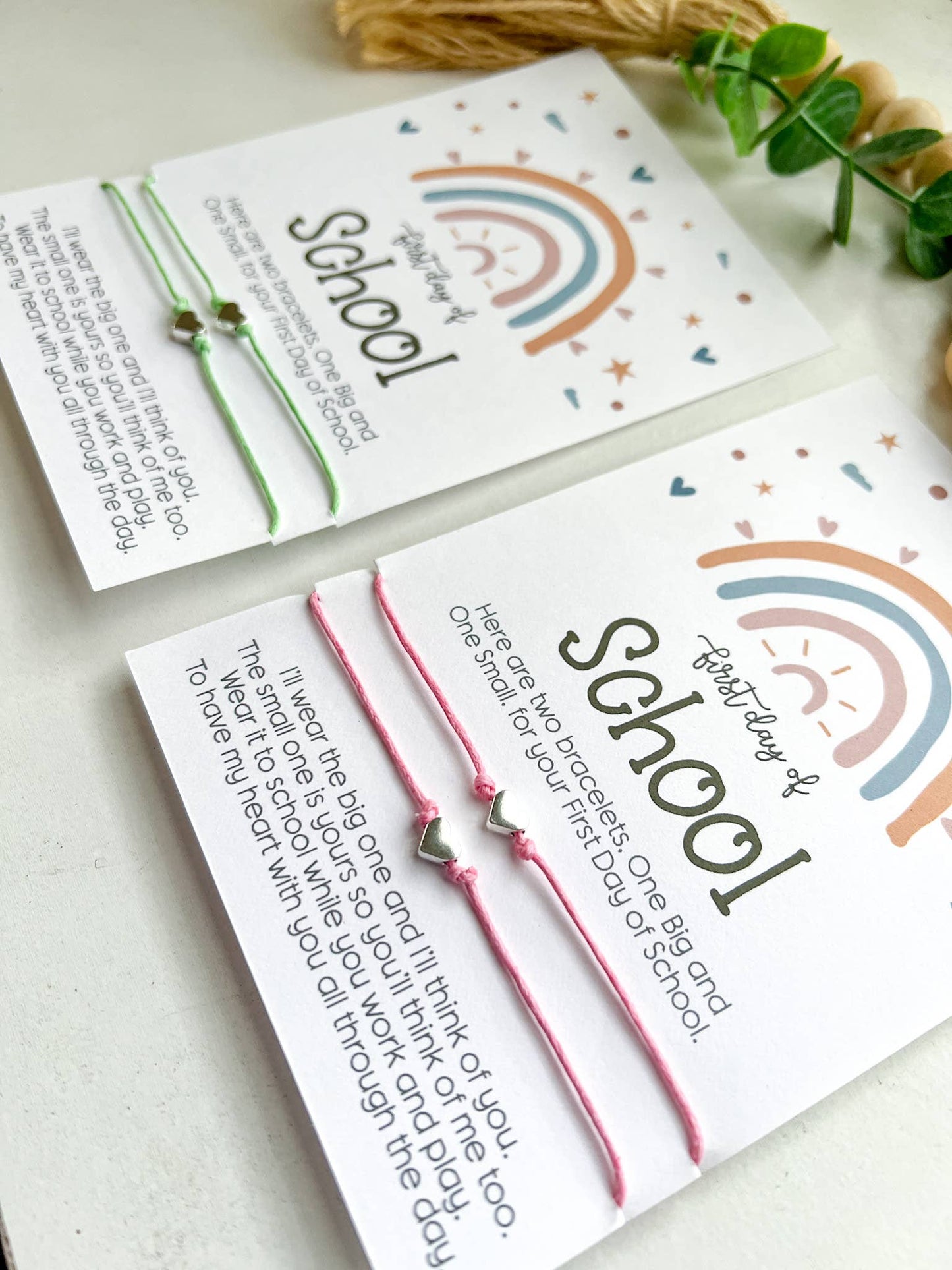 Two "First Day of School" cards featuring a pastel rainbow design with hearts, stars, and clouds. Each card includes a pair of matching string bracelets—one larger and one smaller—with heart-shaped charms in the center. The cards read: "Here are two bracelets. One Big and One Small, for your First Day of School." Below, a heartfelt message explains the bracelets' purpose for connection during the day. One card has mint green bracelets, and the other has pink. Both are placed on a white surface.
