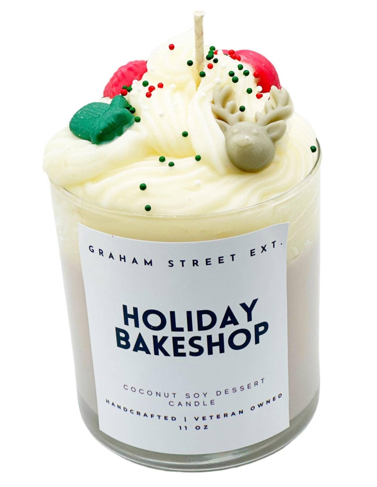 A festive "Holiday Bakeshop" candle crafted with coconut soy wax, presented in an 11 oz glass jar. The candle features a whipped cream-inspired design on top, adorned with colorful holiday decorations including green and red sprinkles, a gray reindeer, and other festive accents, mimicking the look of a holiday dessert. Labeled as handcrafted and veteran-owned by Graham Street Ext.