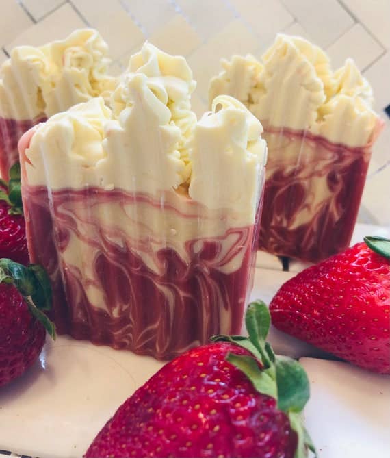 A detailed view of the "Strawberry Shortcake" soap bars showcasing the intricate swirled design of creamy white and berry pink tones, mimicking layers of dessert. The soap is styled alongside plump, ripe strawberries, emphasizing the sweet and fruity theme.