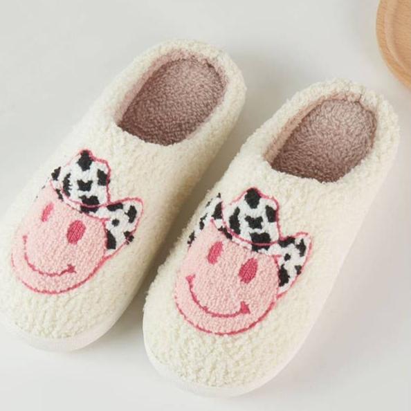 "Cozy cream-colored slippers with a pink smiley face design wearing a black-and-white cow print cowboy hat, displayed on a light background."