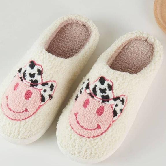 "Cozy cream-colored slippers with a pink smiley face design wearing a black-and-white cow print cowboy hat, displayed on a light background."