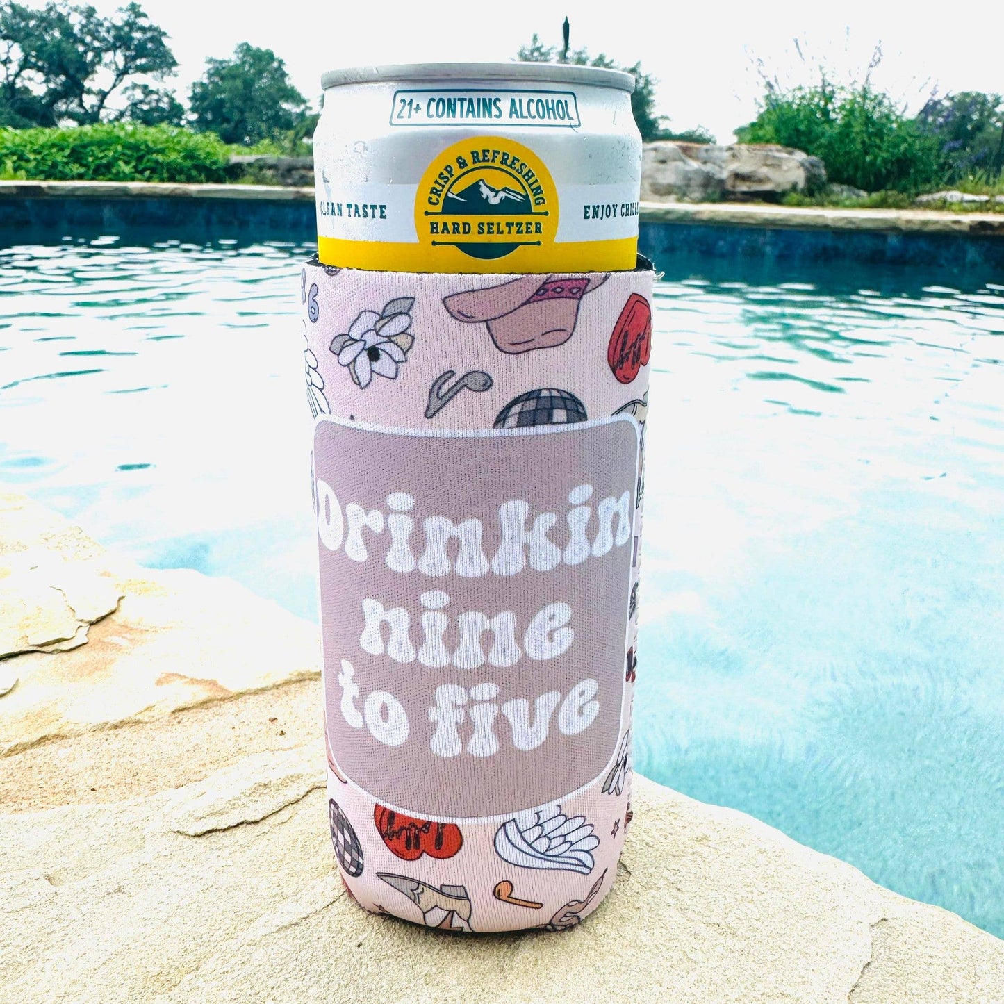 A slim can cooler with a playful design featuring Western-themed illustrations and the text "Drinking nine to five" in bold white letters. The cooler holds a hard seltzer can, placed poolside, creating a fun and laid-back summer vibe.