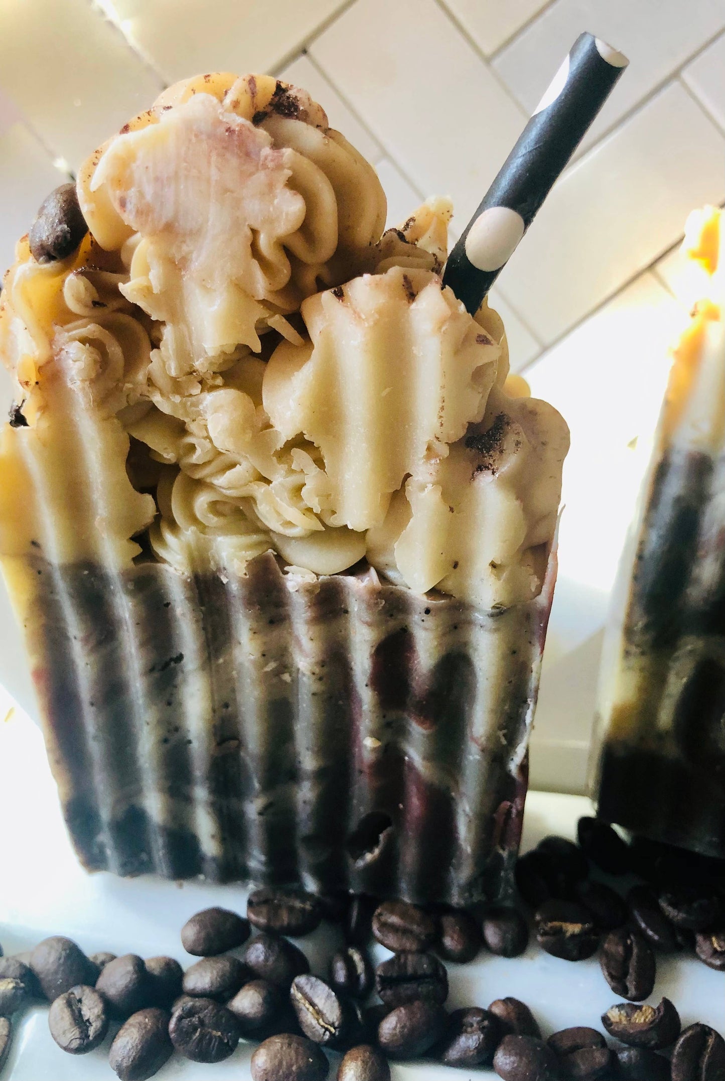 A close-up view of artisan soap bars designed to look like a frothy coffee drink, featuring a textured, creamy beige top resembling whipped cream and a rich brown base mimicking coffee layers. Each bar is adorned with a black-and-white striped straw and sprinkled with coffee grounds, surrounded by scattered coffee beans for a visually appealing, coffee-themed aesthetic.