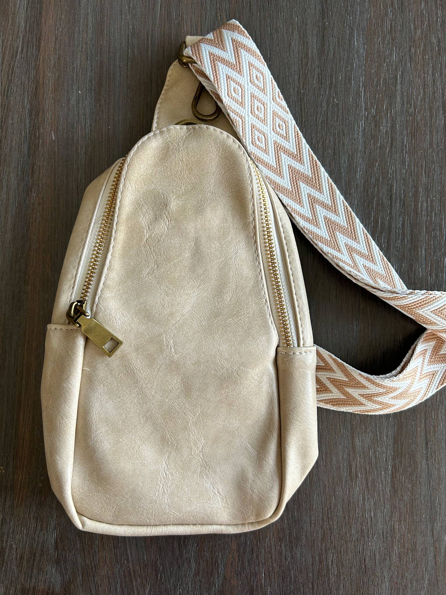 Alt text: A beige faux-leather sling bag with a zipper closure and a patterned strap featuring a tan and white zigzag diamond design. The bag is displayed on a dark wooden surface, showcasing its minimalist and stylish design.
