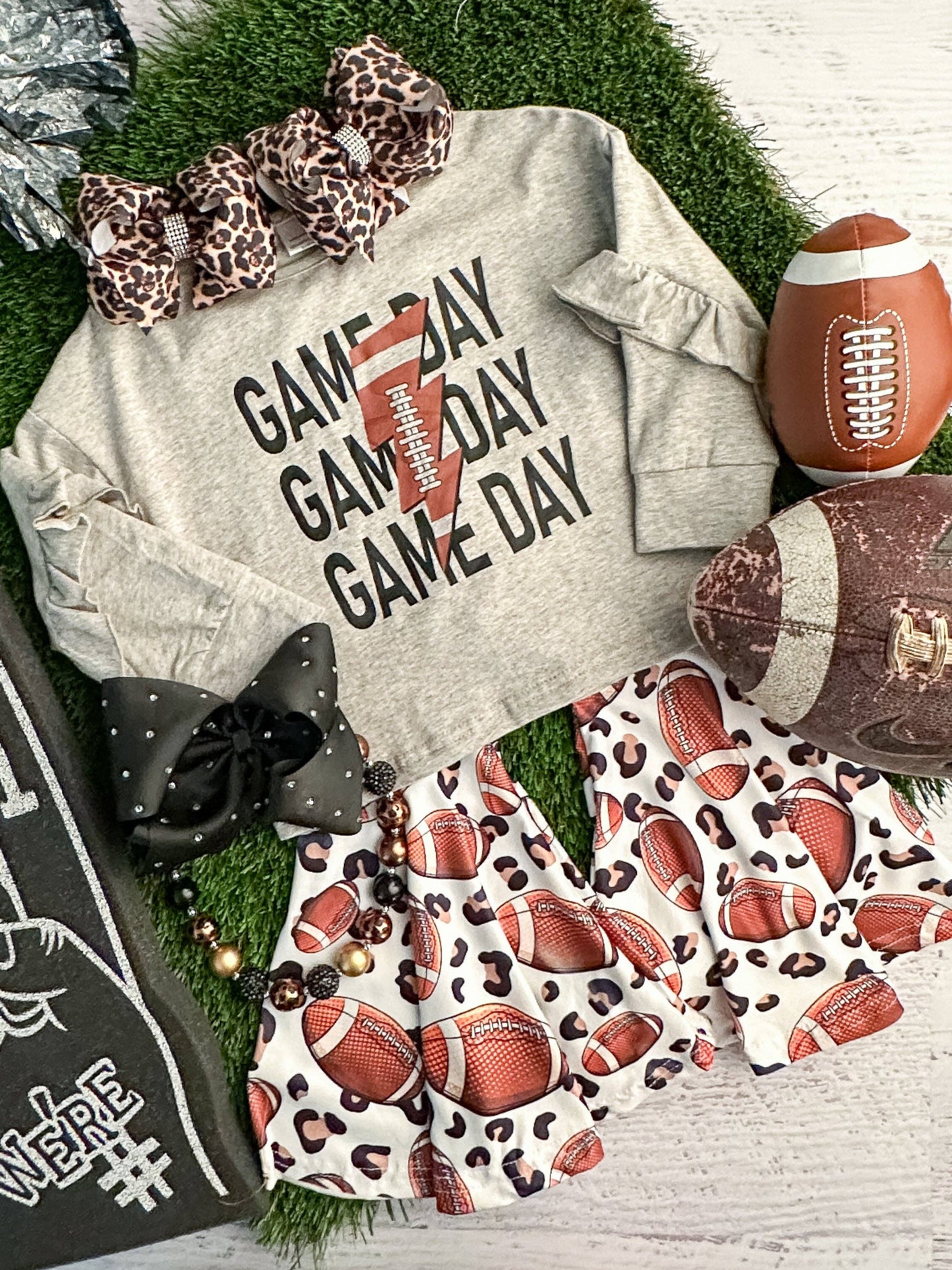 Flat lay of a festive "Game Day" outfit for kids, styled on artificial green turf. The centerpiece is a gray long-sleeve shirt with black text repeating "GAME DAY" and a football graphic with a lightning bolt design. Paired with it are ruffled shorts featuring a pattern of footballs and leopard spots. Accessories include two oversized bows, one in leopard print and the other black with rhinestone accents, as well as a chunky beaded necklace in black, gold, and brown tones. Surrounding props include a brown