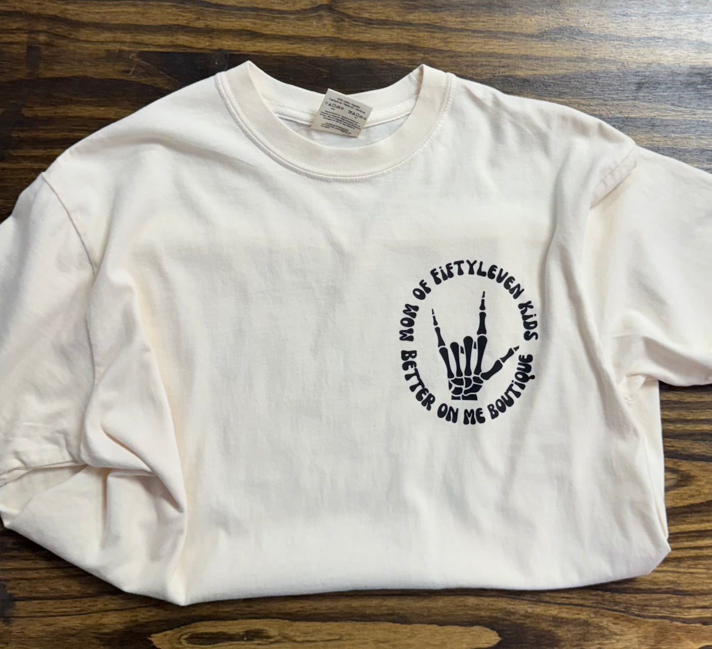 A light cream-colored t-shirt featuring a small, circular graphic on the front left chest. The design includes a skeletal hand making a "rock on" gesture, surrounded by the text "MOM OF FIFTYLEVEN KIDS" and "BETTER ON ME BOUTIQUE" in bold black lettering. The shirt is laid flat on a wooden surface, showcasing its casual and playful vibe.