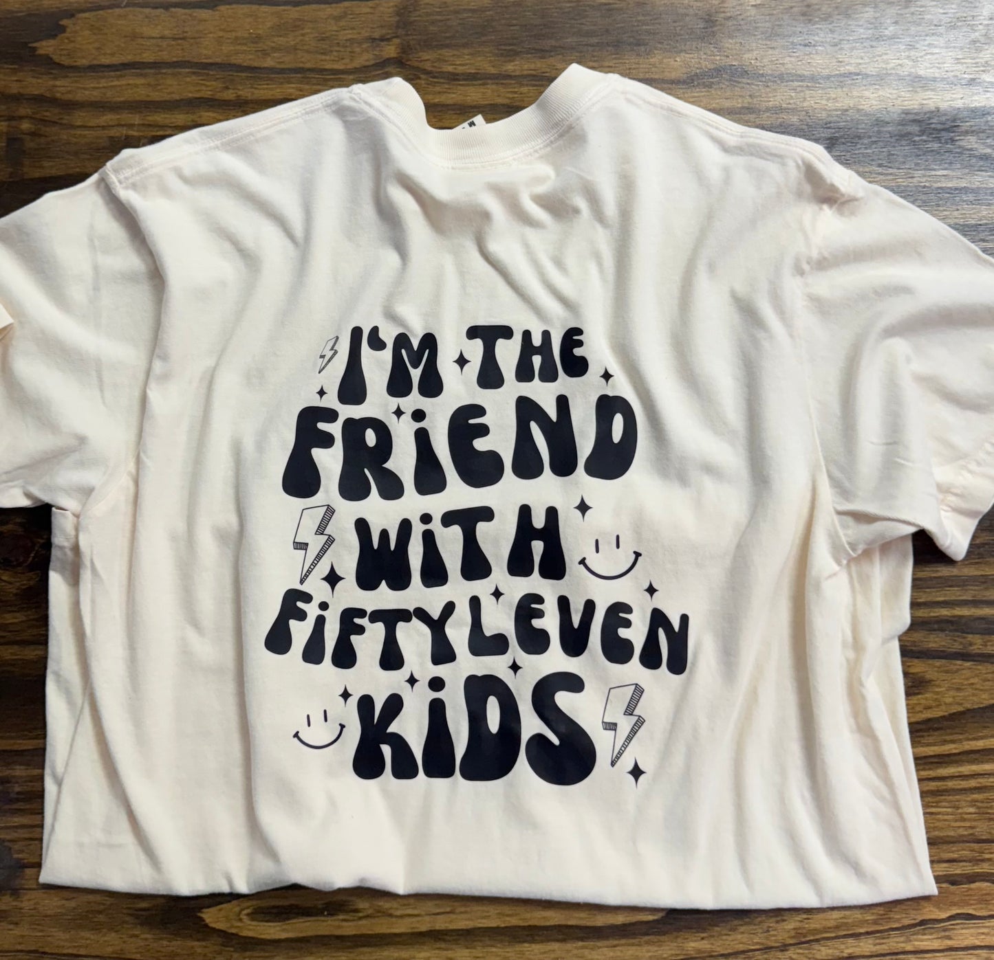 The back of the same t-shirt displays a bold, whimsical black print that reads, "I'M THE FRIEND WITH FIFTYLEVEN KIDS," surrounded by fun doodles like lightning bolts, smiley faces, and star-like sparkles. The design adds humor and personality, perfect for a lighthearted statement. The shirt is again displayed on a wooden surface.
