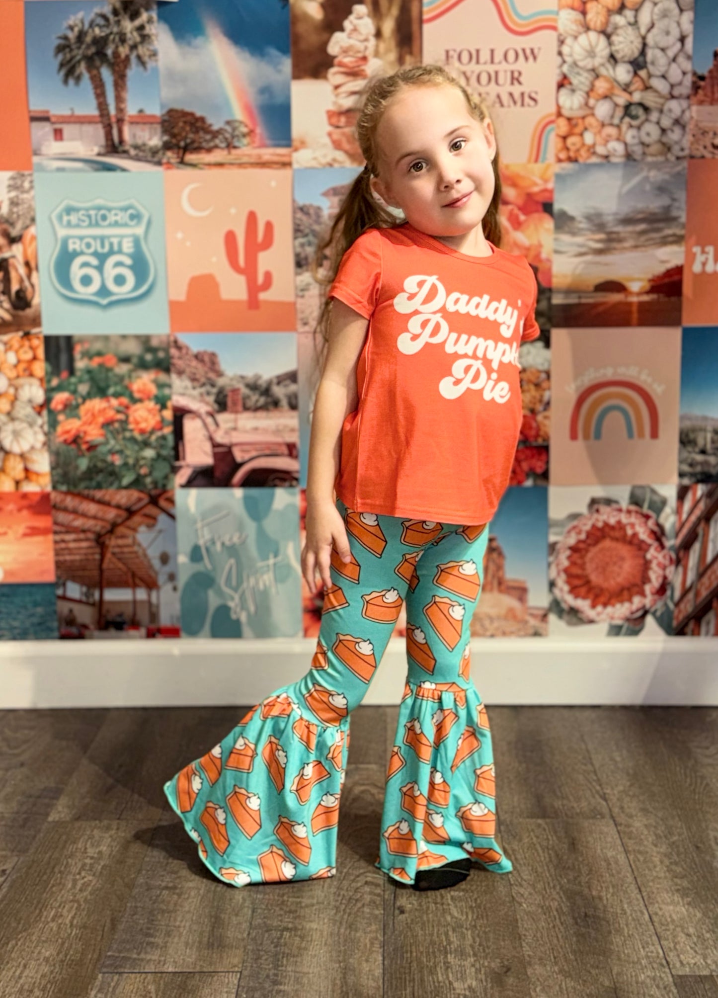 Smiling young girl wearing an orange 'Daddy’s Pumpkin Pie' T-shirt matched with teal bell-bottom pants covered in a whimsical pumpkin pie print. She poses confidently against a colorful backdrop filled with nostalgic Route 66, desert, rainbow, and pumpkin-themed imagery, creating a fun, seasonal vibe.