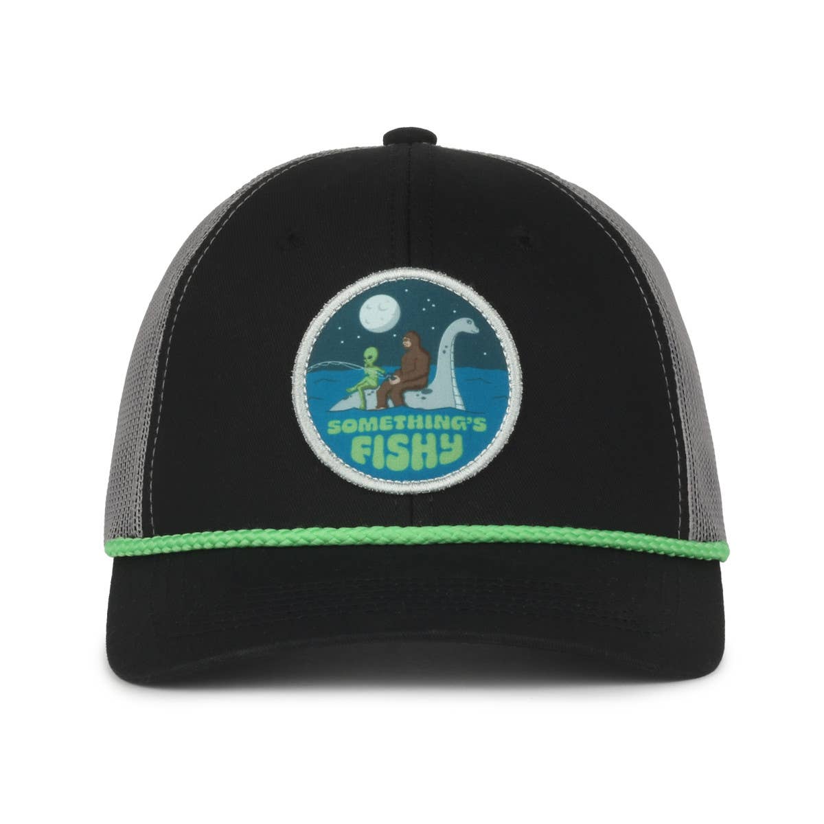 A black and gray trucker hat with a patch featuring a whimsical design of Bigfoot, an alien, and the Loch Ness Monster in a moonlit lake. The words "Something's Fishy" are embroidered in light green below the illustration. The hat has green trim along the front edge.