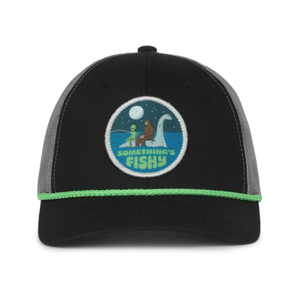 A black and gray trucker hat with a patch featuring a whimsical design of Bigfoot, an alien, and the Loch Ness Monster in a moonlit lake. The words "Something's Fishy" are embroidered in light green below the illustration. The hat has green trim along the front edge.