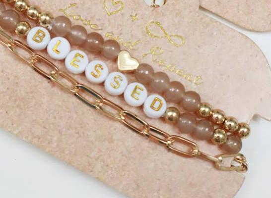 Set of elegant bracelets featuring gold beads, cream beads, and a white letter bead design spelling 'BLESSED.' The set includes a dainty gold heart accent and a stylish gold chain bracelet, displayed on a rustic beige backing card with a minimalist heart design, perfect for gifting or accessorizing with a positive touch.