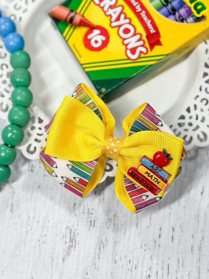 Alt text: A vibrant school-themed hair bow featuring a yellow ribbon layered with a patterned ribbon displaying colorful pencils. The bow is adorned with an enamel charm of stacked books labeled "Art," "Math," and "Reading," topped with a red apple. The bow is displayed on a decorative white plate, accompanied by a green beaded bracelet and a box of Crayola crayons in the background, adding to the back-to-school theme.