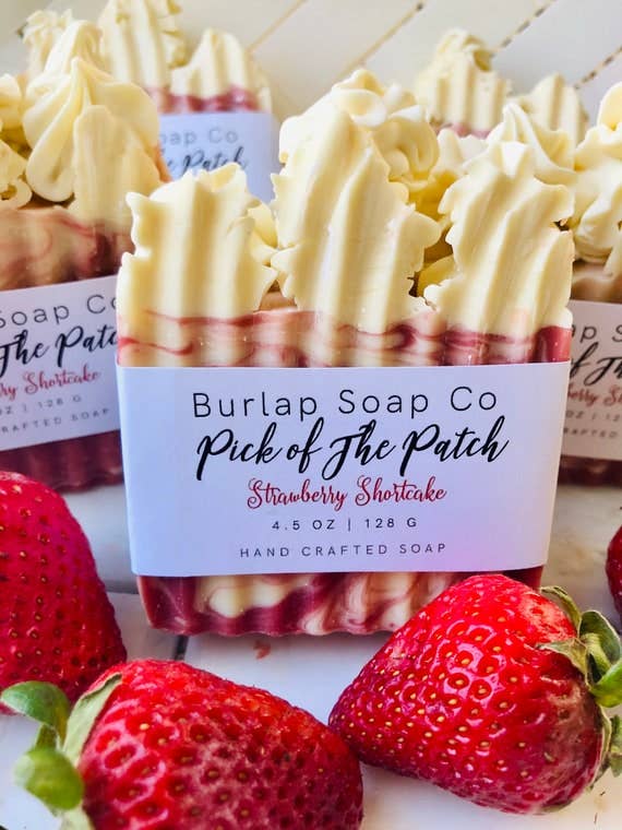 Close-up of handcrafted soap bars labeled "Burlap Soap Co - Pick of the Patch - Strawberry Shortcake." The soap features a swirled design with white and pinkish-red hues, topped with a piped, whipped texture resembling frosting. Fresh strawberries are scattered around the soap, adding a vibrant touch to the image.