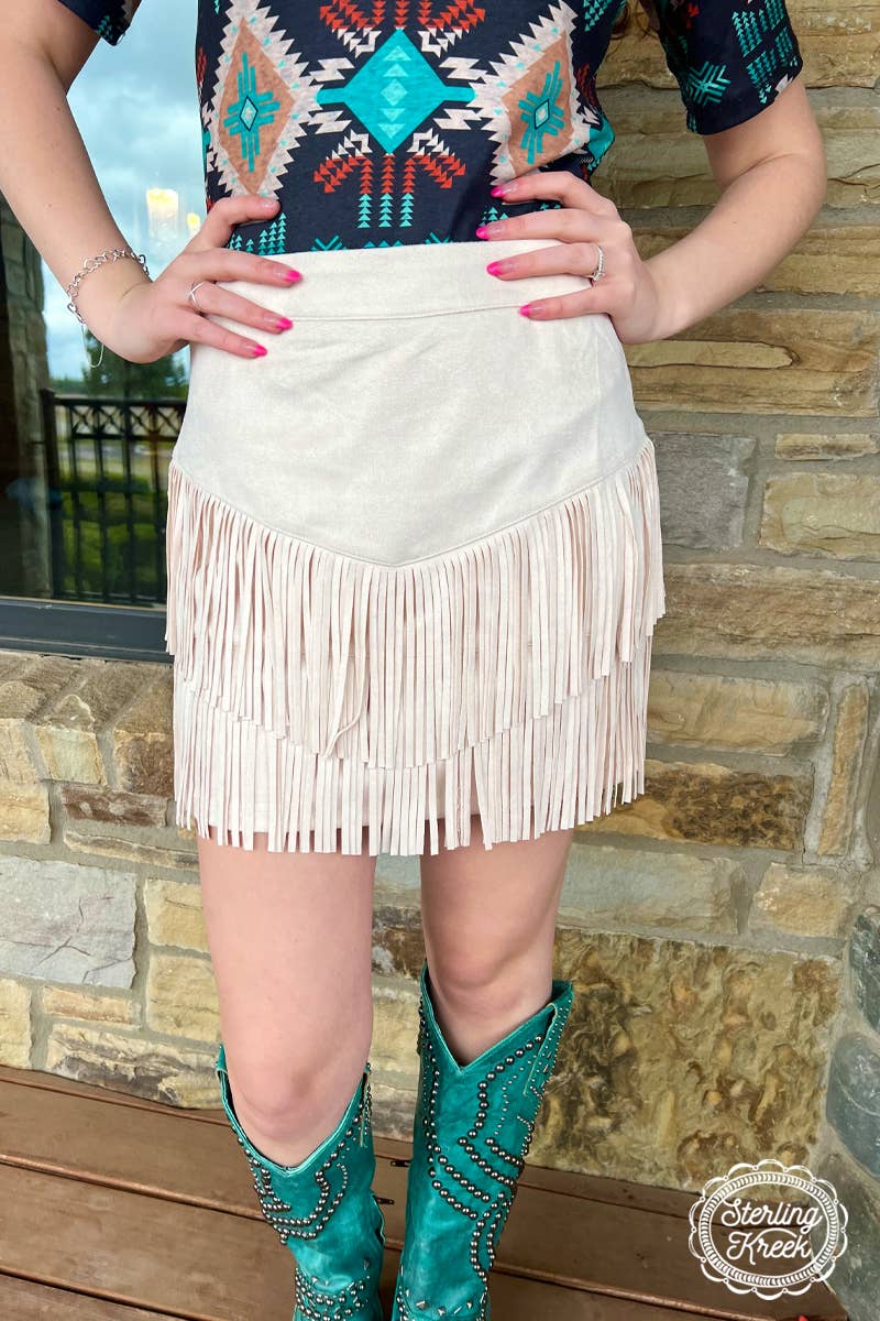 Image of a cream-colored western fringe skirt, featuring a soft, neutral tone that adds a touch of boho-chic to any outfit. The skirt is designed with playful fringe detailing that flows along the hem, creating movement and a relaxed, carefree vibe. The lightweight fabric and flattering fit make it perfect for pairing with boots, sandals, or a tucked-in graphic tee for a laid-back, stylish look.