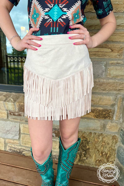 Image of a cream-colored western fringe skirt, featuring a soft, neutral tone that adds a touch of boho-chic to any outfit. The skirt is designed with playful fringe detailing that flows along the hem, creating movement and a relaxed, carefree vibe. The lightweight fabric and flattering fit make it perfect for pairing with boots, sandals, or a tucked-in graphic tee for a laid-back, stylish look.