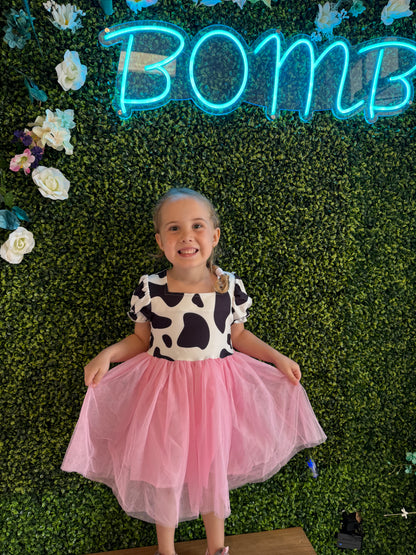 A young girl smiles brightly while wearing a playful dress with a cow-print bodice featuring black spots on a white background and puffed sleeves. The dress has a flowing pink tulle skirt that adds a whimsical touch. She stands barefoot on a wooden platform in front of a green faux foliage wall adorned with white and pastel flowers. Above her, a glowing neon blue sign reads "BOMB," adding a trendy, fun vibe to the setting.