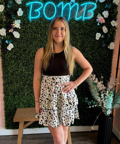 A young woman poses confidently in front of a decorative backdrop featuring a green faux boxwood wall adorned with white and pink flowers, with a glowing neon "BOMB" sign at the top. She wears a sleeveless black top tucked into a white tiered skirt with a black floral pattern, creating a chic and feminine look. Her long, straight blonde hair is styled naturally, and her accessories include a delicate bracelet on her wrist. A wooden stool and a black vase with pastel floral accents are visible in the stylish