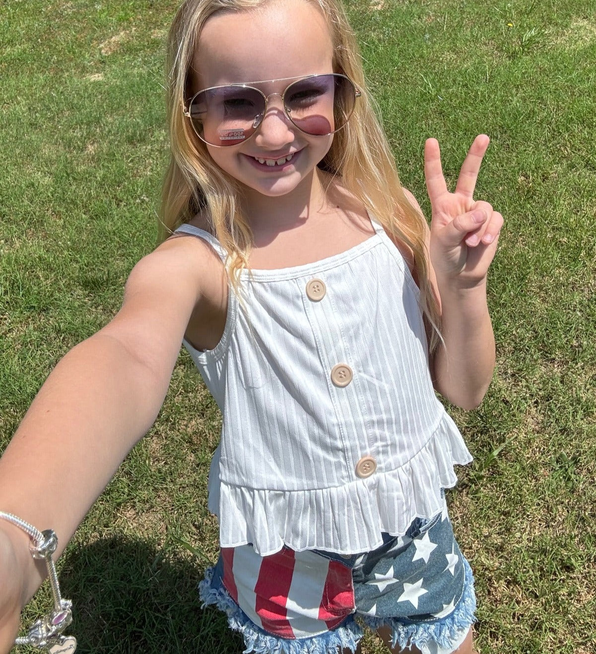 A smiling young girl standing outdoors on grass, wearing aviator sunglasses, a white sleeveless button-up top with a ruffled hem, and distressed denim shorts featuring a patriotic red, white, and blue flag design. She holds up a peace sign, radiating confidence and joy.