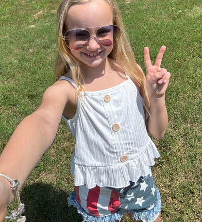 A smiling young girl standing outdoors on grass, wearing aviator sunglasses, a white sleeveless button-up top with a ruffled hem, and distressed denim shorts featuring a patriotic red, white, and blue flag design. She holds up a peace sign, radiating confidence and joy.