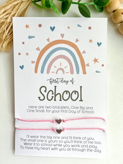 Two "First Day of School" cards featuring a pastel rainbow design with hearts, stars, and clouds. Each card includes a pair of matching string bracelets—one larger and one smaller—with heart-shaped charms in the center. The cards read: "Here are two bracelets. One Big and One Small, for your First Day of School." Below, a heartfelt message explains the bracelets' purpose for connection during the day. One card has mint green bracelets, and the other has pink. Both are placed on a white surface.