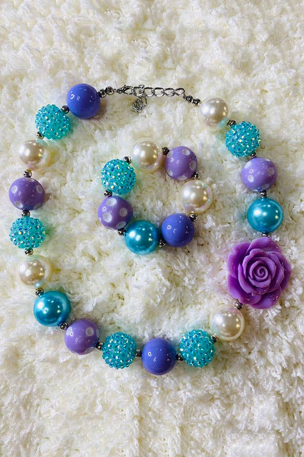 A chunky beaded necklace featuring a mix of purple, turquoise, and pearl beads in various textures and designs, including polka dots, rhinestones, and glossy finishes. The necklace is accented with a large purple rose-shaped bead and is displayed on a soft white textured background.
