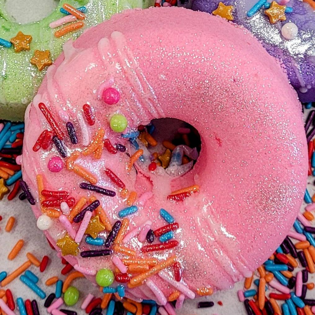 A close-up of a donut-shaped bath bomb in vibrant pink, coated with a sparkling shimmer. The bath bomb is decorated with colorful, candy-like sprinkles, including rainbow jimmies, round beads, and small gold stars, giving it a playful, dessert-inspired look. The texture and detailing make it appear almost edible, creating a whimsical aesthetic for a luxurious bath experience.