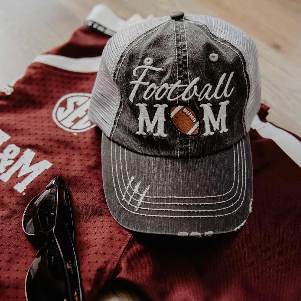 Distressed black and gray trucker hat with white mesh backing, featuring embroidered text "Football Mom" with a football graphic replacing the "O" in "Mom." Styled alongside maroon and white football apparel, making it a perfect accessory for game-day moms supporting their team.