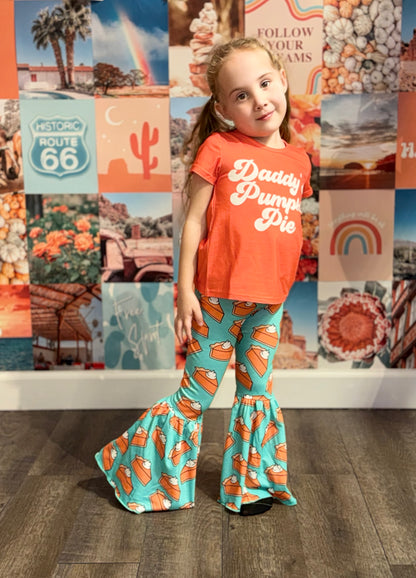 Smiling young girl wearing an orange 'Daddy’s Pumpkin Pie' T-shirt matched with teal bell-bottom pants covered in a whimsical pumpkin pie print. She poses confidently against a colorful backdrop filled with nostalgic Route 66, desert, rainbow, and pumpkin-themed imagery, creating a fun, seasonal vibe.